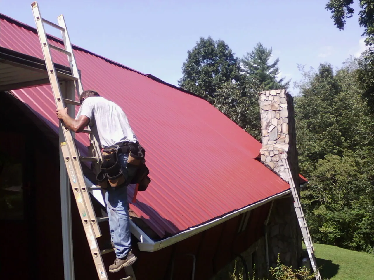 Roofer on a ladder beside a red metal roof and stone chimney — Soffit & Fascia Repair by Roofing Brothers in Veneta